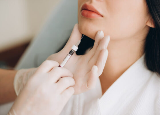 Close up of hands of cosmetologist making botox injection in female lips. She is holding syringe. The young beautiful woman is receiving procedure with enjoyment.